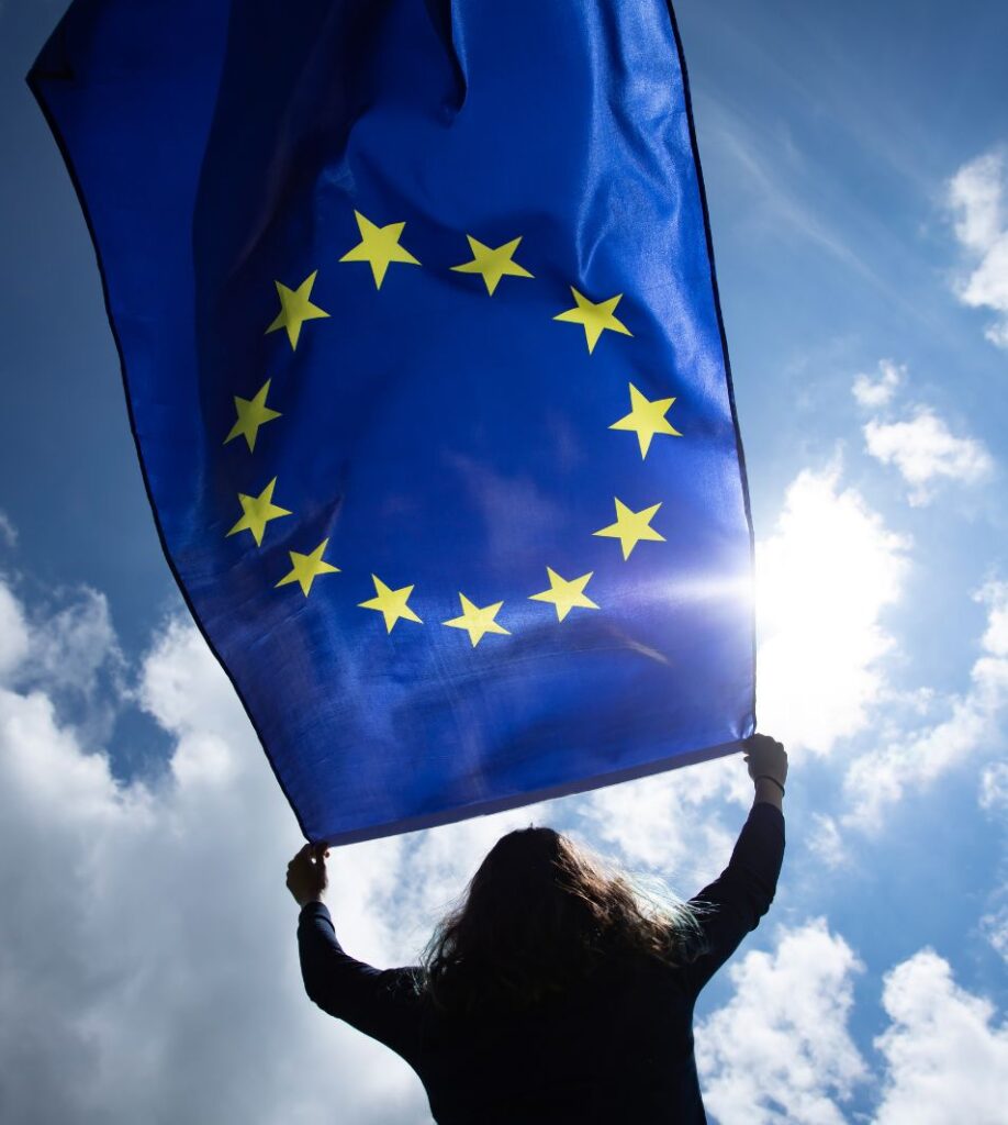 Dłonie trzymające flagę Unii Europejskiej na tle błękitnego nieba.