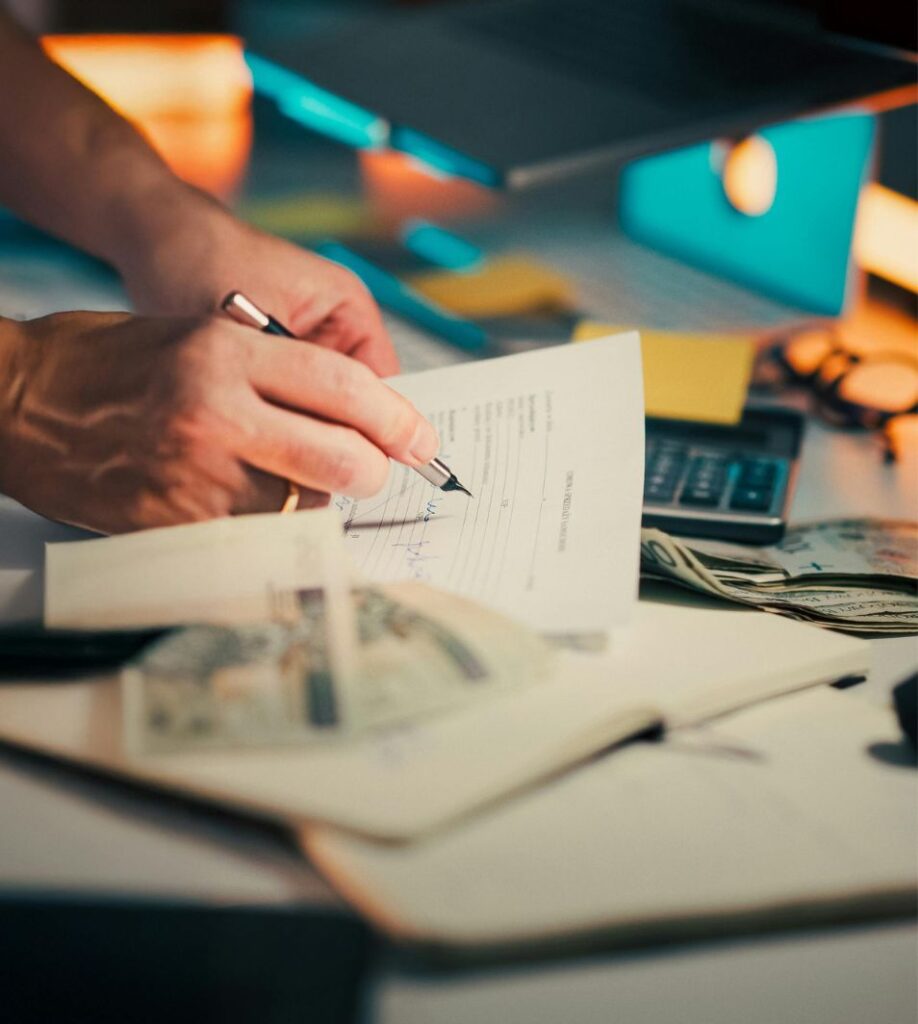 Dłoń podpisująca dokument na biurku obok kalkulatora i banknotów.